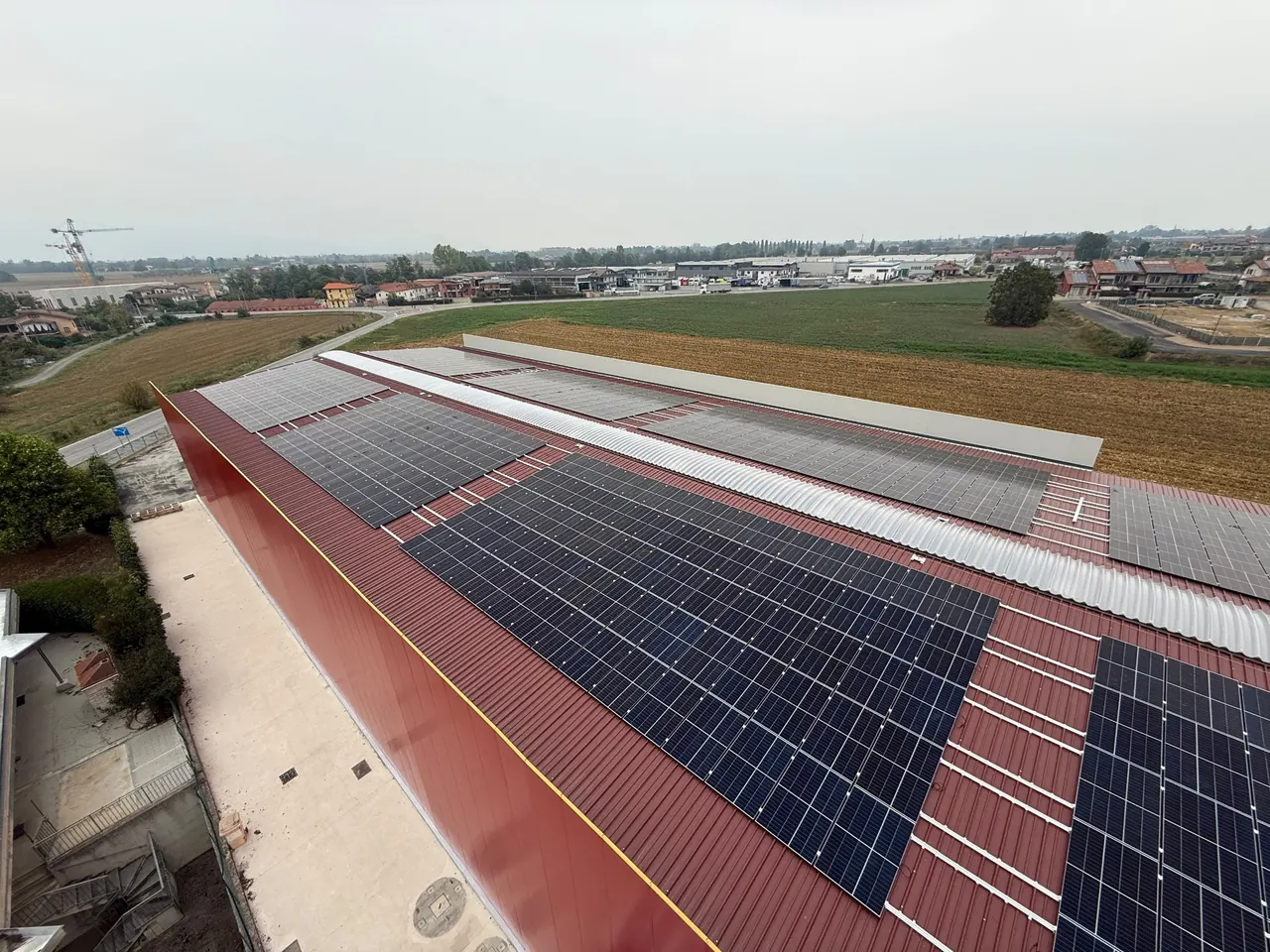 Moduli fotovoltaici su un grande tetto in lamiera rossa visti da un'estremità, con campi coltivati e altri edifici intorno