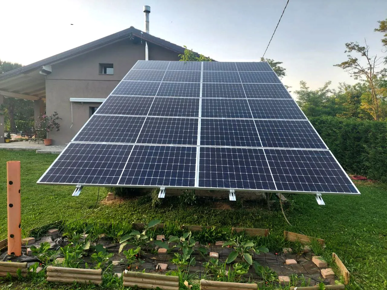 Struttura a terra con moduli fotovoltaici vista da un lato, tra aiuole coltivate e un edificio sullo sfondo