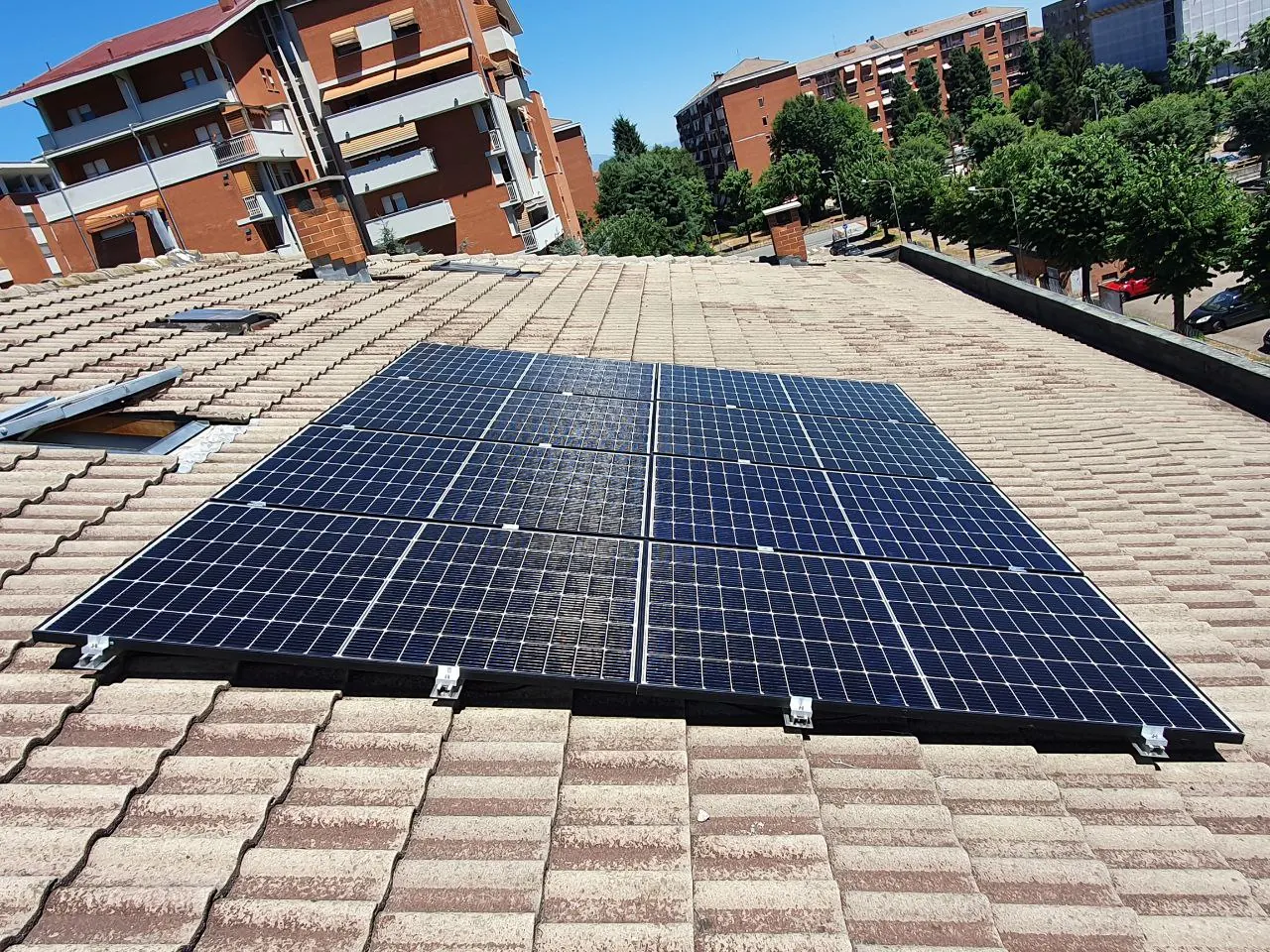 Moduli fotovoltaici su un tetto a falde in tegole, con palazzi residenziali e alberi sullo sfondo