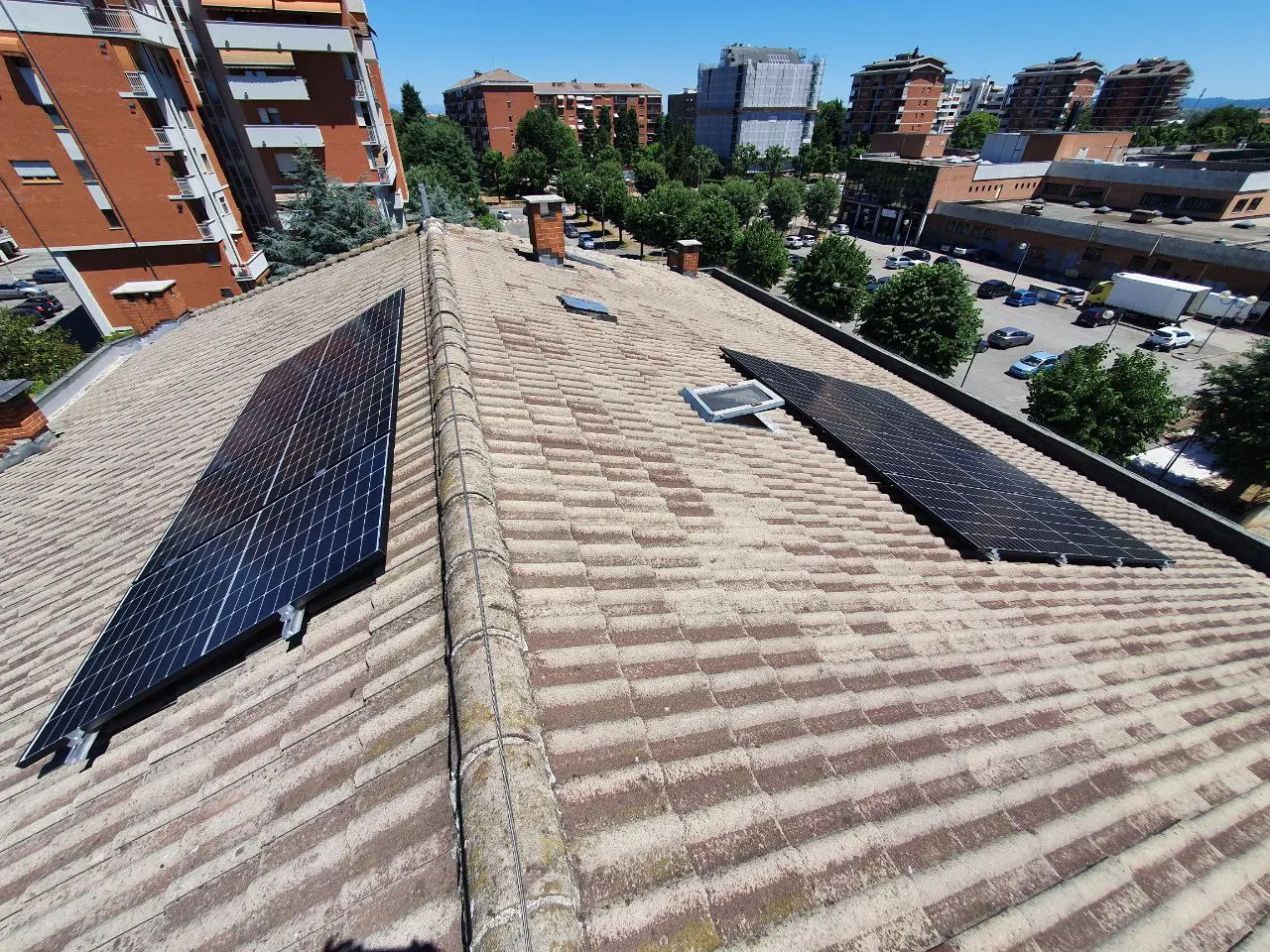 Due gruppi di moduli fotovoltaici su due falde del tetto in tegole, con palazzi e un parcheggio sullo sfondo