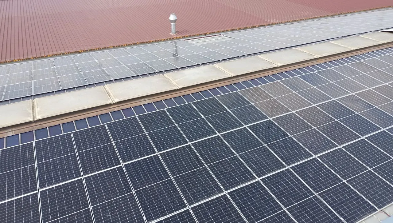 Moduli fotovoltaici in primo piano su un capannone industriale, con la copertura rossa e una fascia chiara sullo sfondo