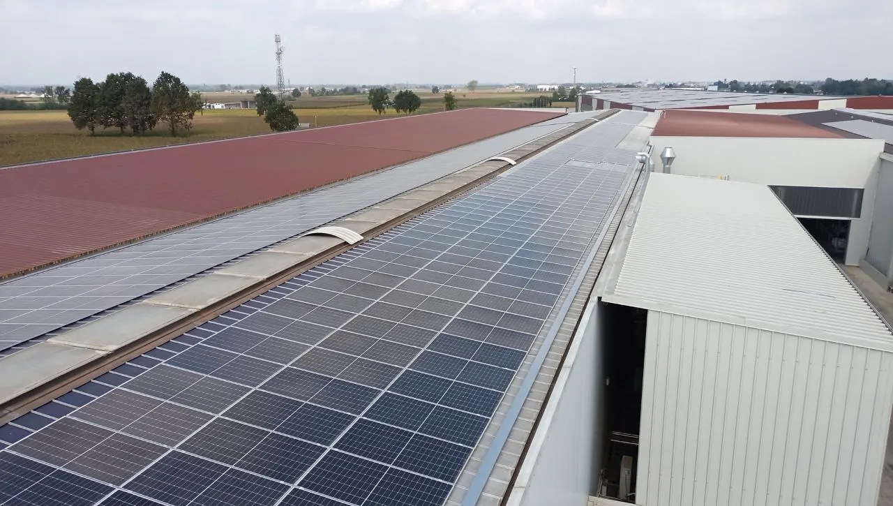 Lunga fila di moduli fotovoltaici lungo la copertura di un capannone industriale, con campi e una torre sullo sfondo