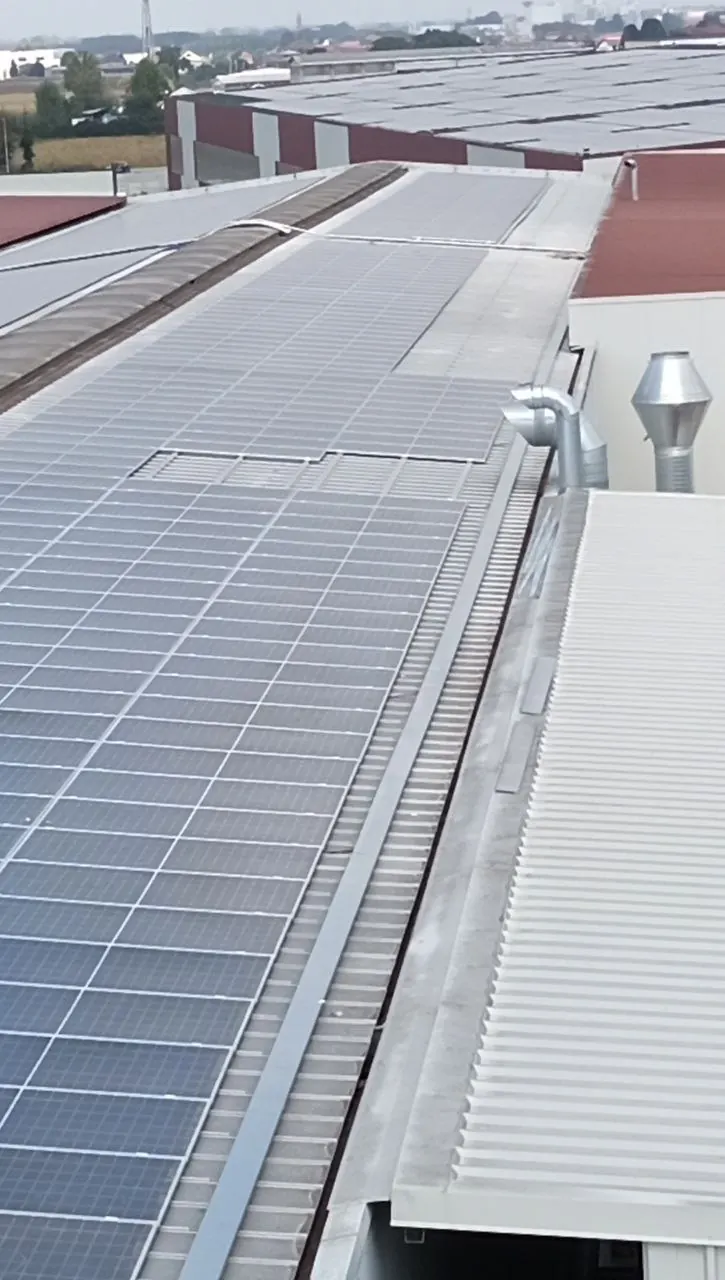 Moduli fotovoltaici lungo la copertura di un capannone industriale, con camini di aerazione e altri edifici sullo sfondo