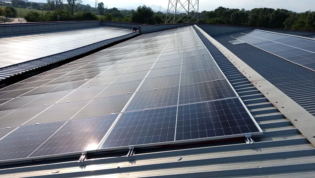 Lunga fila di moduli fotovoltaici lungo la copertura in lamiera grecata, con alberi e un traliccio sullo sfondo