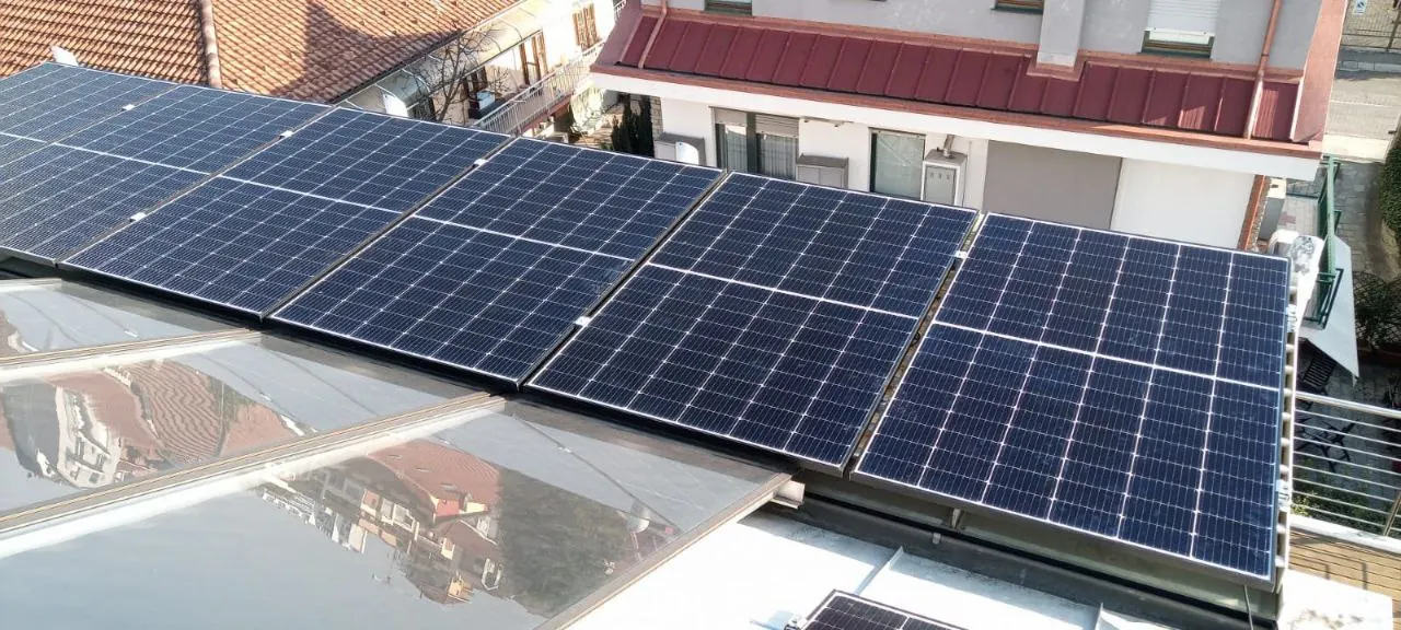 Fila di moduli fotovoltaici accanto a una superficie vetrata, con edifici dal tetto rosso sullo sfondo